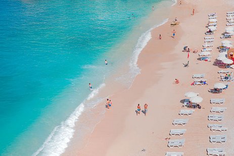 Luftaufnahme eines Strandes mit Menschen und bunten Sonnenschirmen, die sich am Wasser entspannen und die Sonne geniessen.