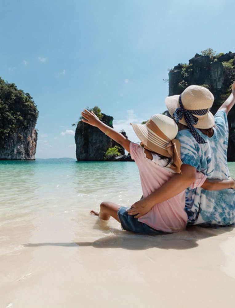 „Asiatische Mutter und Tochter sitzen gemeinsam am Strand und geniessen die schöne Natur während ihrer Ferien. Sommerferien- und Familienreise-Konzept. Reisen in Thailand.