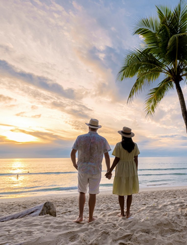 Ein paar Männer und Frauen stehen an einem Palmenbaum und beobachten den Sonnenuntergang am Strand mit weißem Sand und Palmen, Bang Tao Strand Phuket Thailand.
