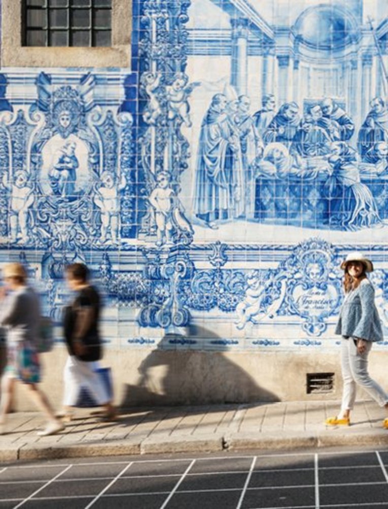 Eine Frau spaziert auf der Straße, während die Sonne scheint und Passanten vorbeigehen.