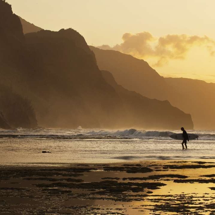 Ein Surfer geht am Strand von Hawai bei Sonnenuntergang entlang, die Wellen im Hintergrund leuchten im warmen Licht.