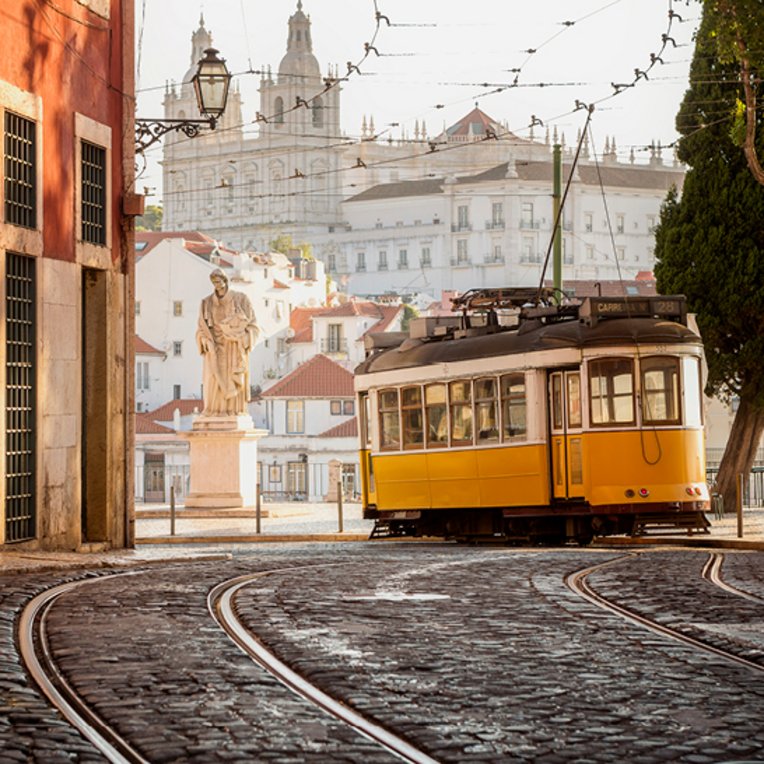 Eine gelb-weisse Strassenbahn fährt über eine Kopfsteinpflasterstrasse in Lissabon.