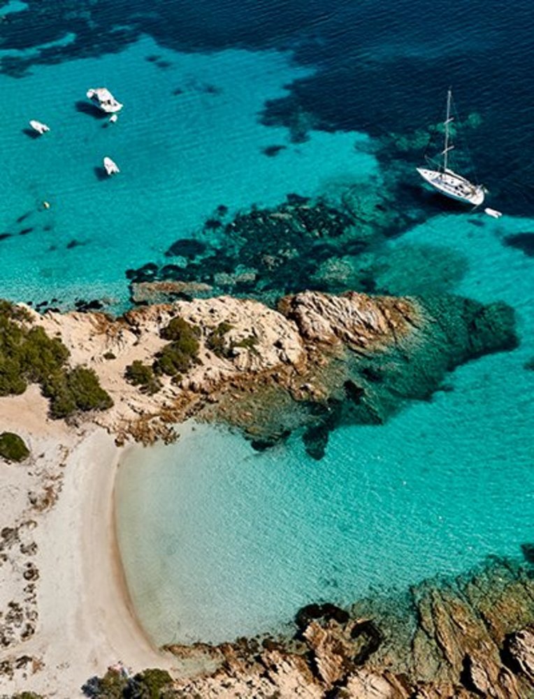 Strand in Sardinien mit Booten im Wasser und einer felsigen Küste im Hintergrund.