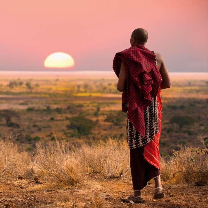 Ein Mann in einem roten Gewand steht bei Sonnenuntergang auf einer Ebene in Tansania.