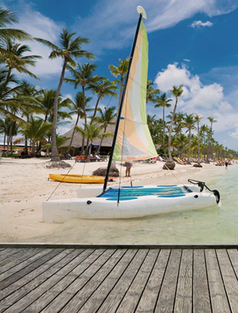 Leere Holzplattform neben einem tropischen Strand in Punta Cana, Dominikanische Republik. Im Hintergrund ein Segelboot, das an der Küste der Sargassosee vor Anker liegt.