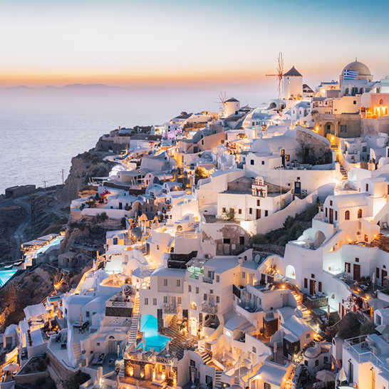 Blick auf die Stadt Oia auf den Kykladeninseln bei Sonnenuntergang, mit leuchtenden Farben am Himmel und ruhigem Wasser.