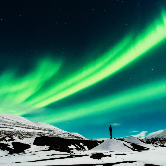 Jemand steht auf einem verschneiten Hügel, während das grüne Nordlicht darüber leuchtet.