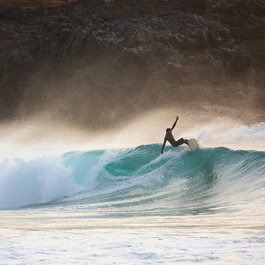 Surfer reitet eine Welle auf Fuerteventura, umgeben von klarem Wasser und strahlendem Himmel.