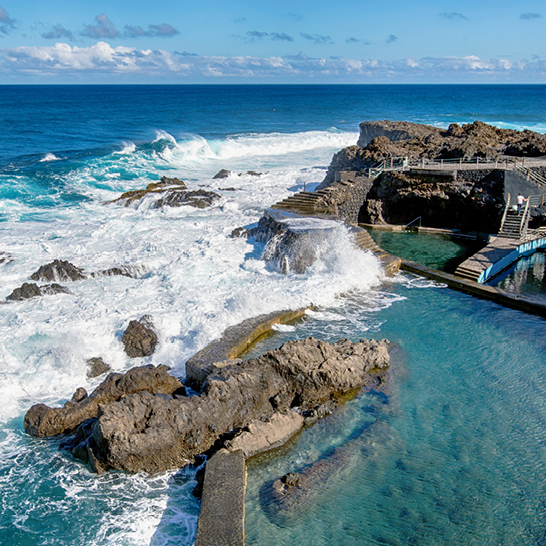 Weitläufiger Pool, umgeben von Felsen und Wasser, mit Palmen, die eine entspannte Urlaubsstimmung vermitteln.