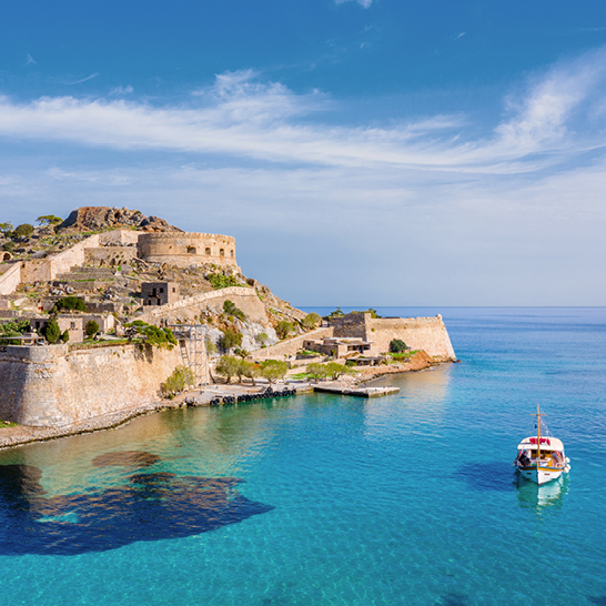  Ein Boot treibt im Wasser, während das Schloss Spinalonga im Hintergrund sichtbar ist.