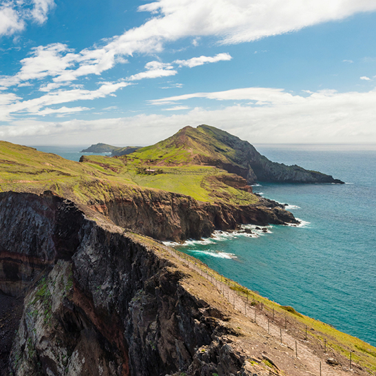 Blick von einer Klippe auf den Ozean, mit Wellen, die gegen die Felsen schlagen und einem klaren blauen Himmel.