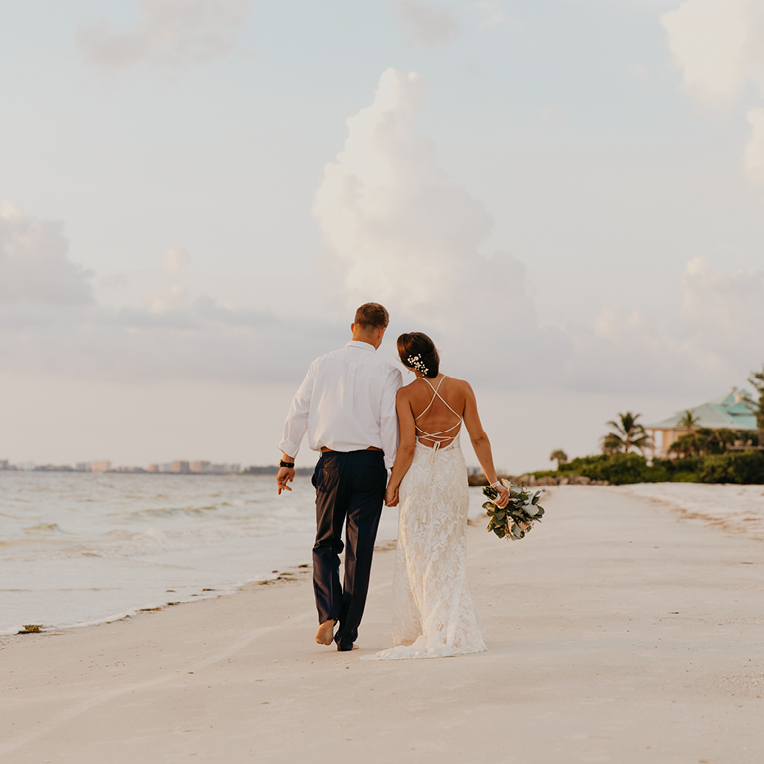 Jungvermählte, die Hand in Hand am Strand spazieren, umgeben vom Klang der Wellen und sanftem Sonnenuntergangslicht.